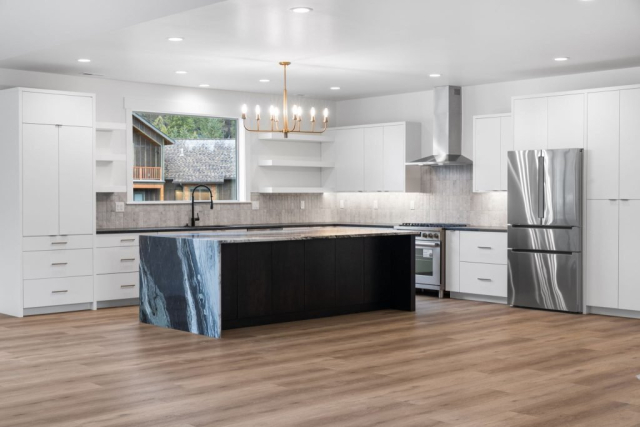 A custom modern kitchen with white cabinets, stainless steel appliances, and a large island. The island features a striking black and white marble countertop. A gold chandelier hangs overhead, while light wood flooring complements the room. A window offers a view of trees and buildings.