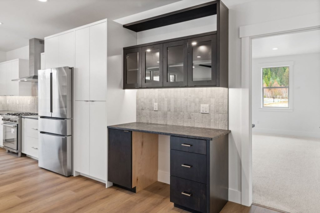 A modern, spacious kitchen features sleek white cabinetry, a high-quality stainless steel refrigerator, and a gas stove with an overhead range hood. A built-in dark wood desk area with glass-front upper cabinets is tastefully integrated into the kitchen. A doorway opens to a carpeted room.