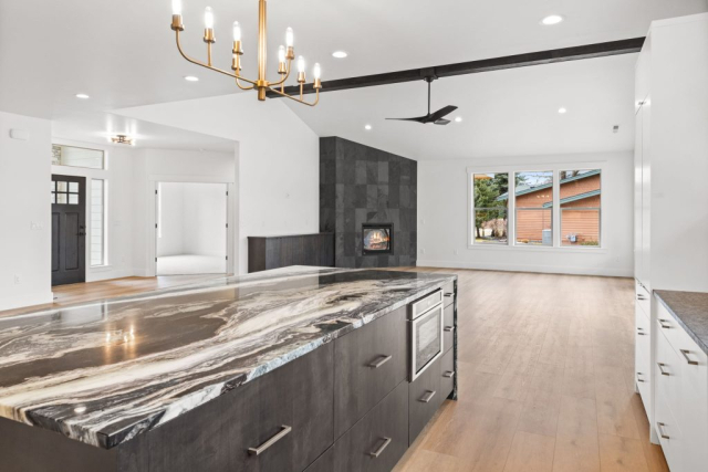Spacious modern kitchen and living area with an open floor plan in this custom home. The kitchen features a large marble island, sleek cabinets, and stainless steel appliances. The living area includes a clean black tiled fireplace, wooden floors, large windows, and a modern chandelier.