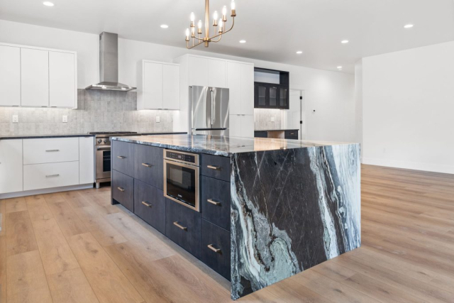A sleek, modern kitchen with wooden flooring features a large island with a black and white marbled waterfall countertop. The clean design includes built-in appliances like an oven and drawers. White cabinets, stainless steel appliances, and a designer chandelier complete the space.