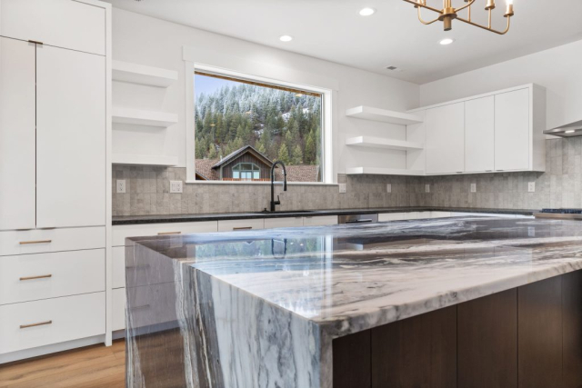 Modern, spacious kitchen with a marble-topped island and dark wood base. Features white cabinets, light gray backsplash, and a large window revealing a scenic view of a house and forested hillside. A sleek black faucet adorns the sink, and a quality gold chandelier hangs overhead.