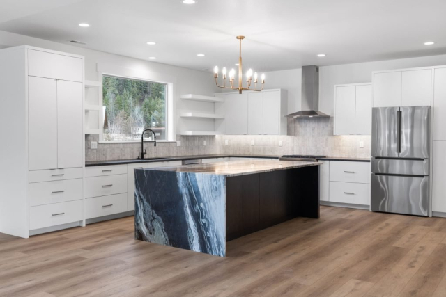 A spacious, modern kitchen with white cabinets, stainless steel appliances, and a light gray backsplash. The center island features a black base with a striking white and blue marble countertop. A gold chandelier hangs above, and a large window provides natural light for an exceptionally clean look.