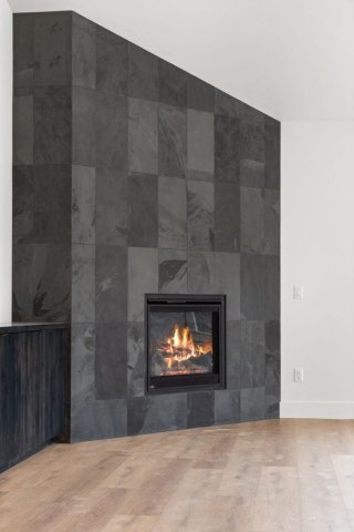 24-web-or-mls-92-marie-victoria-ct A spacious, minimalist living room features a sleek, modern fireplace with a black tiled surround. A cozy fire burns in the hearth, casting a warm glow on the light wooden floor. The walls are painted white, highlighting the clean contrast with the dark fireplace tiles.