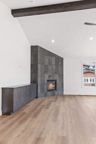 25-web-or-mls-92-marie-victoria-ct A spacious, minimalist living room features a sleek, modern fireplace with a black tiled surround. A cozy fire burns in the hearth, casting a warm glow on the light wooden floor. The walls are painted white, highlighting the clean contrast with the dark fireplace tiles.