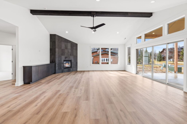 27-web-or-mls-92-marie-victoria-ct A modern living room in this custom home features a sleek, black-tiled corner fireplace with a small, built-in wooden cabinet to the left. The room boasts high, vaulted ceilings with exposed wooden beams, hardwood flooring, and white walls. A large window allows natural light to fill the quality space.