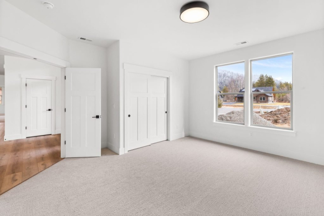 31-web-or-mls-92-marie-victoria-ct A bright, clean room with white walls and large windows letting in natural light. The floor is carpeted in a light color. There are two white doors, one open leading to a hallway with wooden flooring and the other to a closet. A designer ceiling light fixture is visible, adding a touch of elegance to this custom home space.