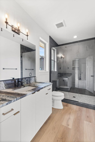 33-web-or-mls-92-marie-victoria-ct A modern bathroom in this custom home features a dual-sink white vanity, black marble countertop, and large mirror. To the right is a glass-enclosed shower with gray tiles and a white toilet. The light wood floor enhances the clean design, illuminated by natural light and wall-mounted lights.
