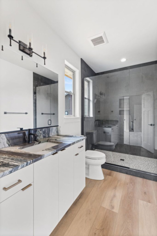 34-web-or-mls-92-marie-victoria-ct A modern bathroom in this custom home features a dual-sink white vanity, black marble countertop, and large mirror. To the right is a glass-enclosed shower with gray tiles and a white toilet. The light wood floor enhances the clean design, illuminated by natural light and wall-mounted lights.