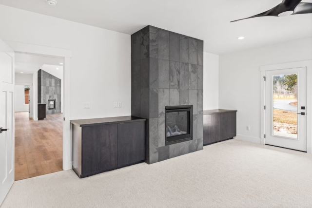 42-web-or-mls-92-marie-victoria-ct A modern living room with a sleek black tiled fireplace, built-in dark wood cabinets on either side, white walls, and a ceiling fan. This designer space features a door to the outside and an open entryway leading to a room with quality wooden flooring.