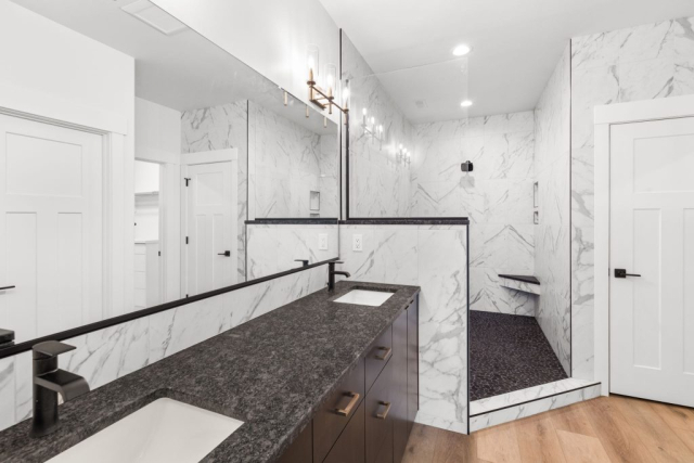 44-web-or-mls-92-marie-victoria-ct A spacious modern bathroom features a black granite countertop with double sinks, a large wall mirror, and sleek black fixtures. The walk-in shower, enclosed by marble-patterned tiles, includes a bench and a black showerhead. The light wood floor and white doors complete this custom home's elegant design.