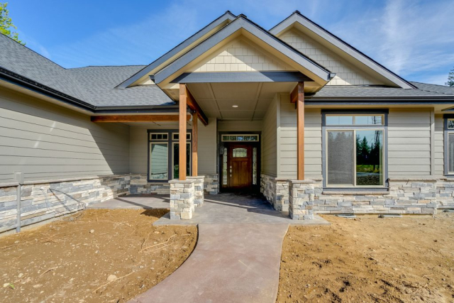 A newly constructed, spacious two-story custom-designed residential home featuring a blend of wooden siding and stone accents, with multiple gables and a covered entryway, set against a backdrop of tall evergreen trees