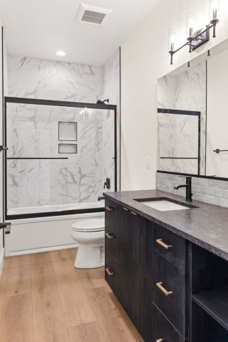 51-web-or-mls-92-marie-victoria-ct A modern bathroom featuring a natural wood floor, a spacious black vanity with a white sink and black faucet, and a large mirror under a sconce light fixture. The clean bathtub has a glass door and marble-patterned tile surround with built-in shelves, reflecting designer elegance.