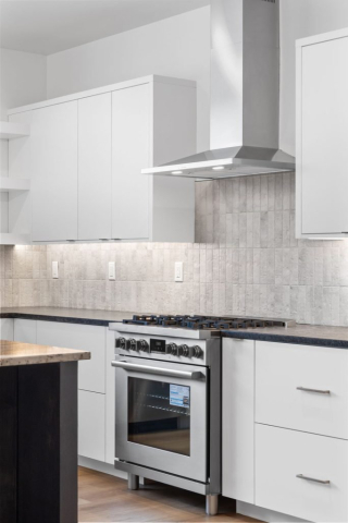 Modern kitchen with white cabinets, a stainless steel range hood, and a gas stove. The backsplash features light-colored, textured tiles. The countertops are dark, providing contrast to the white cabinets and light walls. This designer kitchen in a custom home boasts a spacious wooden island.