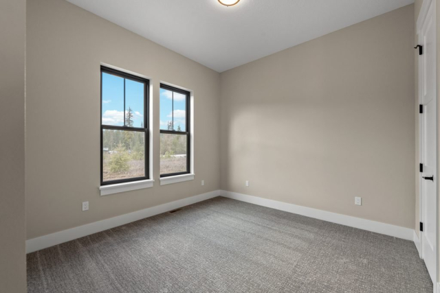 2111 E Garwood Rd A small, modern room with beige walls and a gray carpet. Two tall windows on one wall offer an outside view. The spotless space has white baseboards and trim, with a closed white door on one side. Sunlight streams through the windows, lighting up the spacious room.