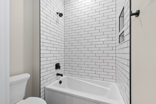 2111 E Garwood Rd A modern bathroom featuring a white bathtub and shower combination exudes designer quality. The shower walls are lined with clean, white subway tiles, and black fixtures are present, including a showerhead and faucet. A built-in alcove for toiletries is visible within the shower wall.
