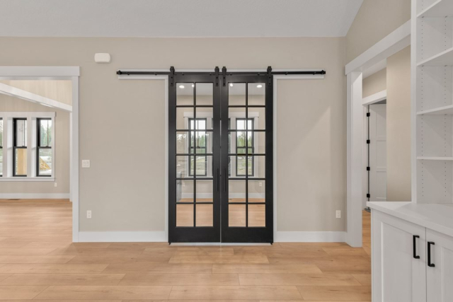 2111 E Garwood Rd A modern, spacious living space features light wood flooring, beige walls, and a black sliding barn door with glass panels. Adjacent rooms and windows with black frames are visible. White baseboards and built-in shelving contribute to the clean, minimalist look designed for contemporary living.