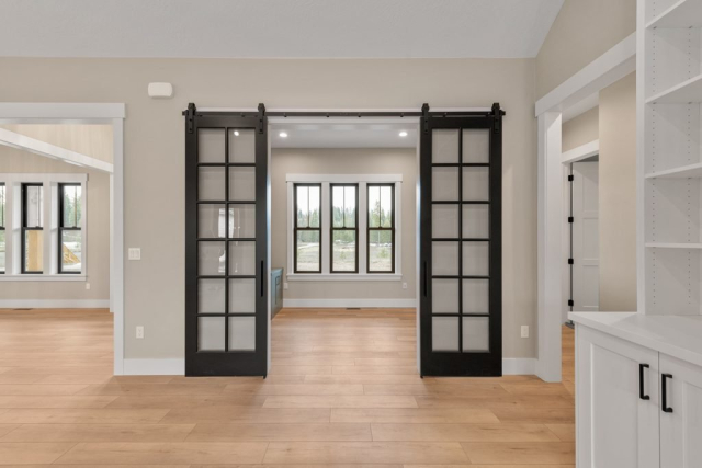 2111 E Garwood Rd A modern, spacious living space features light wood flooring, beige walls, and a black sliding barn door with glass panels. Adjacent rooms and windows with black frames are visible. White baseboards and built-in shelving contribute to the clean, minimalist look designed for contemporary living.