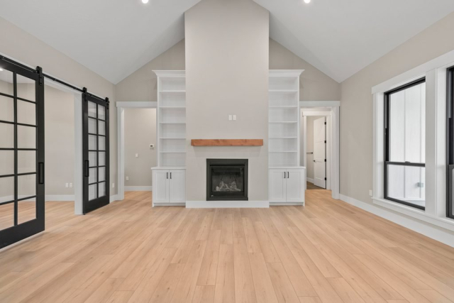 2111 E Garwood Rd A spacious, empty living room with light wood flooring and high vaulted ceilings. This quality, custom home features a centrally located fireplace flanked by built-in shelves. Black-framed sliding glass doors are on the left, and large windows on the right. The gray walls add a modern touch.