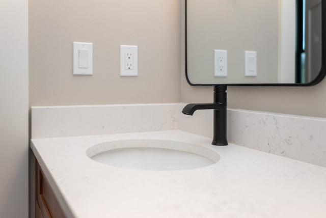 2111 E Garwood Rd A modern bathroom sink with a round white basin set in a white marble countertop. Above the sink is a wall-mounted mirror with a black frame, flanked by dual white electrical outlets. The sleek black faucet on the right of the basin adds to the clean, quality finish of this custom home design.