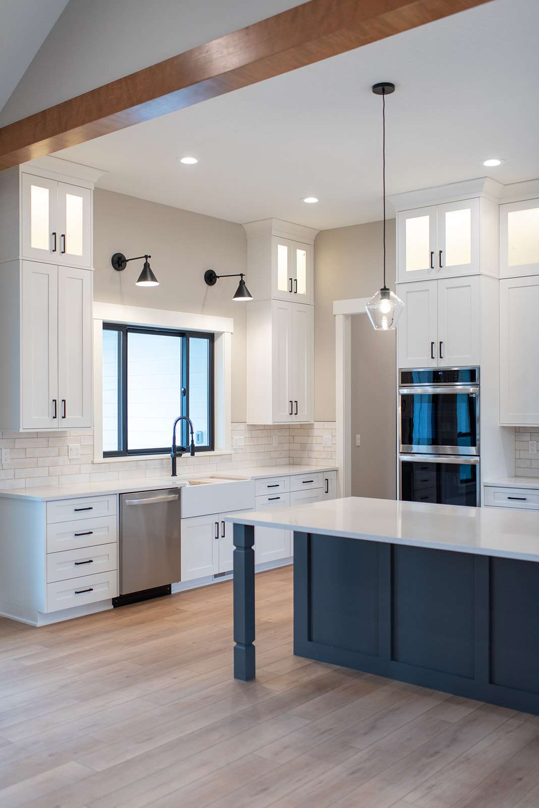 2111 E Garwood Rd A modern kitchen in a custom home featuring white cabinets with black handles, stainless steel appliances, a black faucet, and light-gray subway tile backsplash. The island has a dark base with a light countertop, and two pendant lights hang overhead. Light wooden flooring adds to the spacious feel throughout.