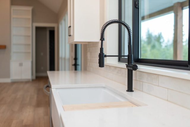 2111 E Garwood Rd A modern kitchen in a custom home features a sleek, white countertop with an integrated sink. A black, industrial-style faucet with a flexible hose is installed above the sink. A large window with a view of greenery outside is positioned above the sink, allowing ample natural light into the spacious area.