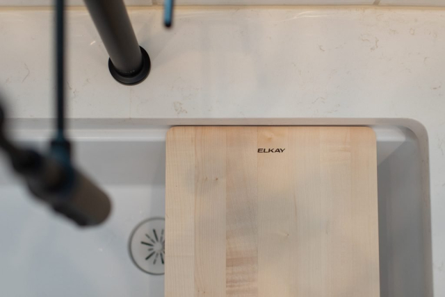 2111 E Garwood Rd A close-up of a kitchen sink in a spacious custom home, with a wooden cutting board partially covering it. The cutting board has the word "ELKAY" engraved on it. The edge of the faucet and a portion of the quality countertop are visible.