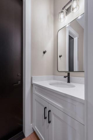 2111 E Garwood Rd A designer bathroom features a modern white countertop and sink, complemented by a sleek black faucet. Quality white cabinetry with black handles contrasts beautifully with the rectangular mirror framed in black. Two exposed lightbulbs sit above the mirror, while a dark wooden door stands adjacent.