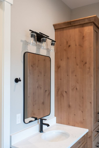 2111 E Garwood Rd A modern bathroom features a quality rectangular mirror with black trim, a designer black faucet over a white countertop and sink, and a three-light wall fixture with glass shades above the mirror. A wooden cabinet with a natural finish stands adjacent to the sink area.