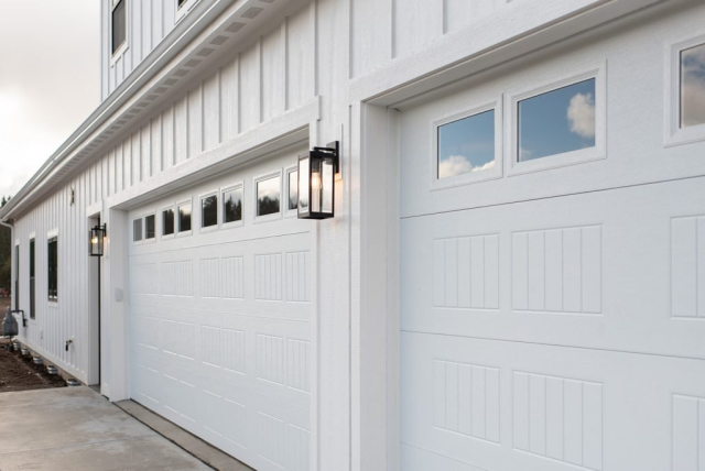 2111 E Garwood Rd A close-up image of a designer modern white house with two large garage doors. Each door features rectangular windows at the top. The clean exterior wall is textured, and two decorative black lantern-style lights are mounted between the garage doors. The driveway is concrete.