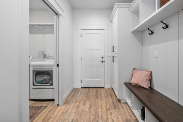 E Lisa Rd by Premier Quality Homes A modern laundry room with a custom built washer and dryer on the left, a wooden bench with a pink cushion on the right, complimented by wall hooks and open shelving above.