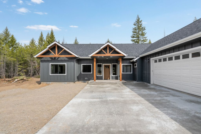 A new custom-built modern single-story house with gray siding and stone accents, featuring a large garage and an inviting entrance with wooden beam details, set against a backdrop of pine trees under a clear blue sky