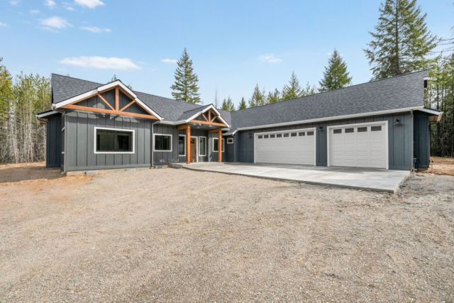 Modern single-story house with gray siding and stone accents, designed featuring a large three-car garage and a well-maintained gravel driveway, set against a backdrop of pine trees under a clear blue sky.