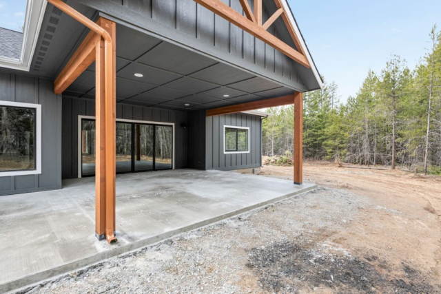 Modern home exterior featuring a spacious concrete patio with stylish wooden beams, nestled in a tranquil, natural wooded setting. This new residence is the epitome of serene living.