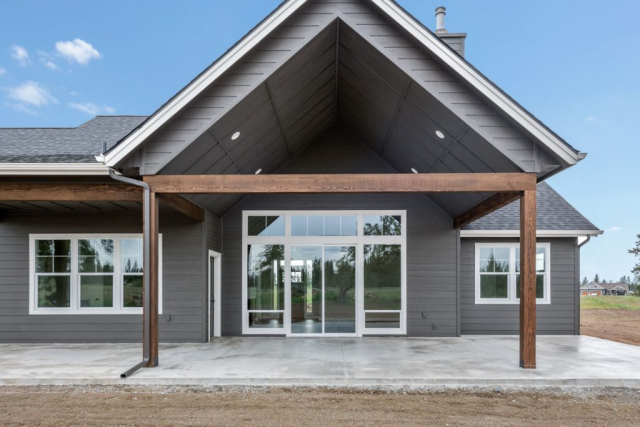 Modern home exterior with a spacious patio, featuring a custom-built gable roof design with wooden beams, large sliding glass doors, and a sleek gray finish.