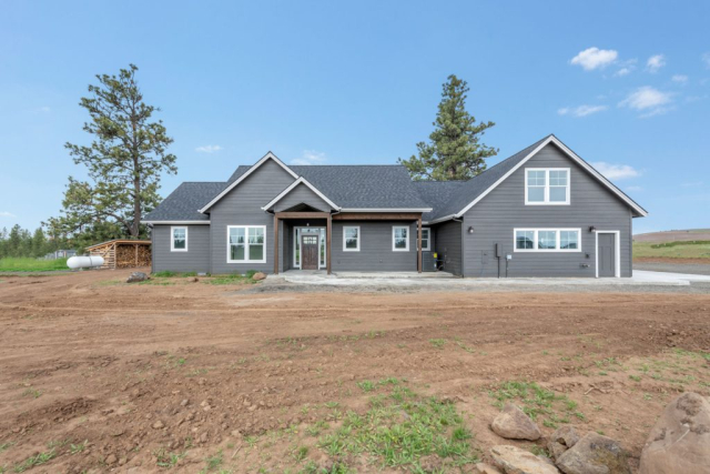 A newly constructed single-story gray house with a dark roof, set against a clear blue sky and surrounded by a barren yard waiting for landscaping, is a custom-built home designed to impress.
