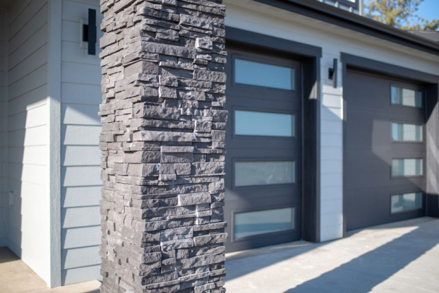 Modern home exterior designed featuring a textured stone pillar with grey accents, complementing the sleek gray garage doors and clean architectural lines.