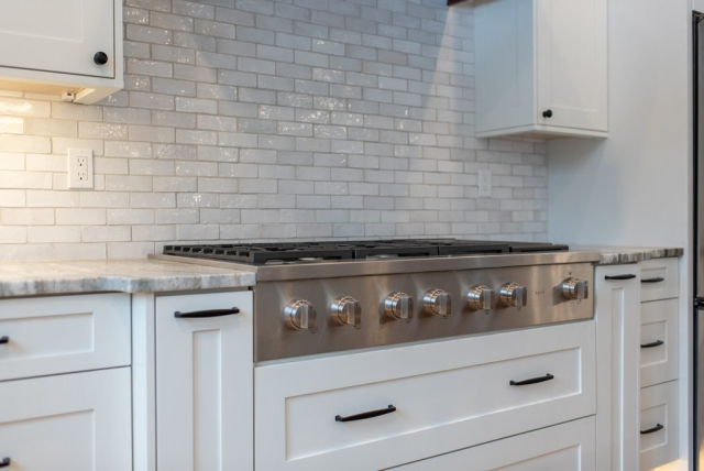 25362 N Werre Rd A modern kitchen with a stainless steel gas stove featuring six burners. The clean backsplash consists of glossy white rectangular tiles. The quality white cabinetry has black handles, and the countertop is a light-colored stone.