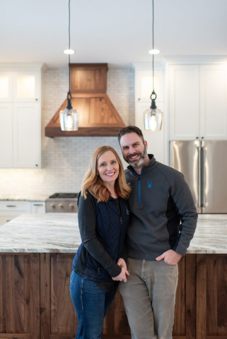 25362 N Werre Rd A smiling couple stands in a spacious, modern kitchen with white cabinets, stainless steel appliances, and a marble countertop. Wooden accents, including a hood vent and the island, add warmth. Two pendant lights hang over the island in this clean, custom home, enhancing the welcoming atmosphere.