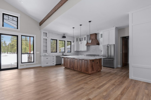 25362 N Werre Rd A clean, spacious, modern kitchen with high ceilings, wood accents, and large windows. It features a wooden beam, a central island with a sink and pendant lights above, stainless steel appliances, white cabinetry, and hardwood floors. A designer door opens to an outdoor area.