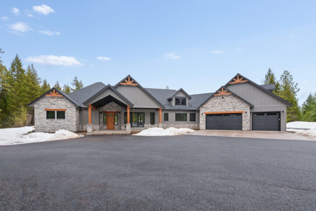 25362 N Werre Rd A spacious stone and wood house with a dark gray roof features dormer windows and an attached three-car garage. The quality design includes an asphalt driveway, snow patches, and trees in the background under a clear blue sky.