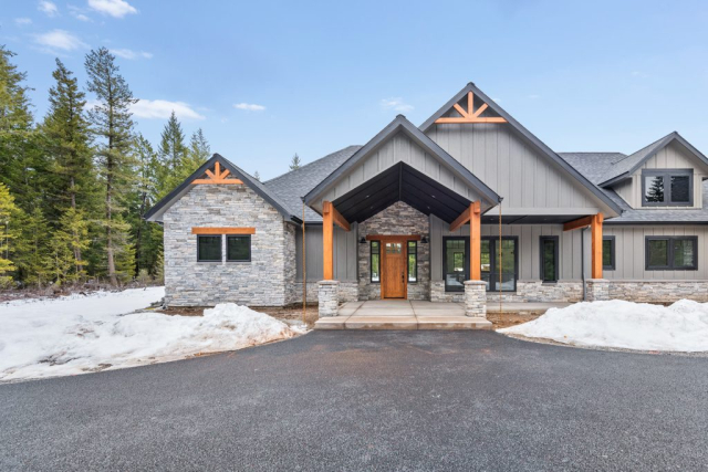 25362 N Werre Rd A modern, rustic-style custom home with a stone and wood exterior is set against a backdrop of a forest. The front features a peaked roof, a covered porch, and a wooden door. Snow is piled on both sides of the path leading to the entrance.
