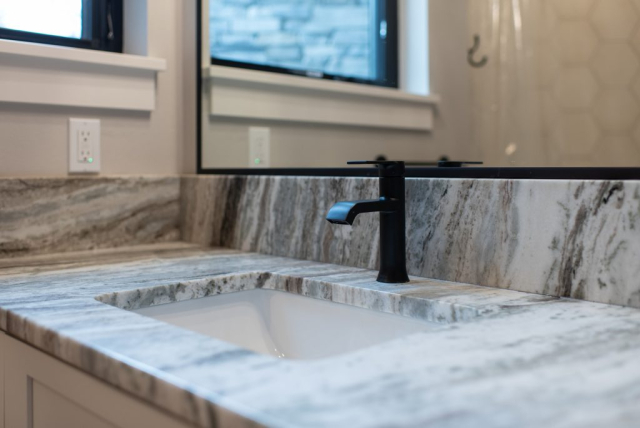 25362 N Werre Rd A modern bathroom vanity with a marble countertop, a rectangular under-mount sink, and a black faucet in a custom home. A large mirror with a white frame is mounted on the wall above the vanity, adding to the clean look. The background shows a window and a glimpse of a hexagonal-tiled wall, creating a spacious feel.