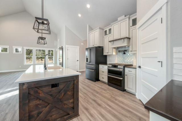 Modern kitchen interior designed for custom homes, featuring white cabinetry, stainless steel appliances, a center island with a unique wooden base, and sleek hardwood flooring, all bathed in natural light.