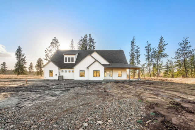 Newly constructed, custom-built home in a rural setting with a gravel terrain in the foreground and sparse trees under a clear sky.