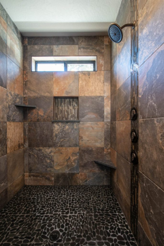 Woodlake Drive by Premiere Quality Homes A modern shower with slate tile walls, pebble floor, and metallic fixtures in custom homes, featuring a built-in shelf and a small window for natural light.