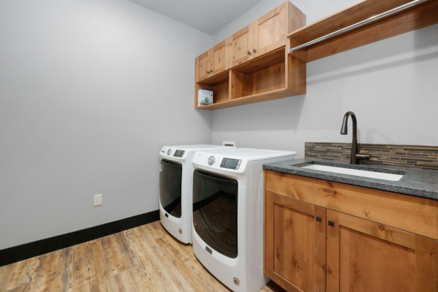 Woodlake Drive by Premiere Quality Homes A modern laundry room with custom-built wooden cabinets, a washing machine and dryer set, and a utility sink with a gray countertop against a neutral wall color.