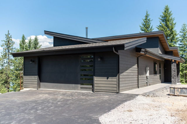 Modern single-story custom-built house with a dark facade and angled roofline, featuring a large garage door with horizontal windows, set against a backdrop of green trees under a clear blue sky.