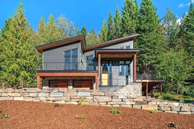 parade-house-1 Modern two-story house with large windows, wood and stone accents, and a sloped roof. It is surrounded by a lush green lawn and tall trees under a clear blue sky. A set of stairs leads up to the main entrance, and a gravel path borders the grass.