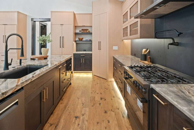 parade-house-14 Modern kitchen with wooden floor, light wood cabinets, and dark lower cabinets. An island features a marble countertop with a potted plant. Built-in shelves display books and decor, and warm lighting creates an inviting atmosphere.