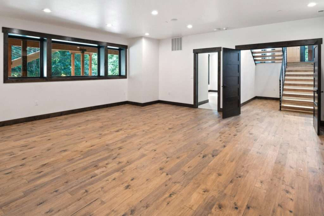 parade-house-31 A spacious room with light wood flooring and white walls features large windows with dark frames. An open dark wood door leads to a staircase. Natural light illuminates the room, creating a bright and airy atmosphere.